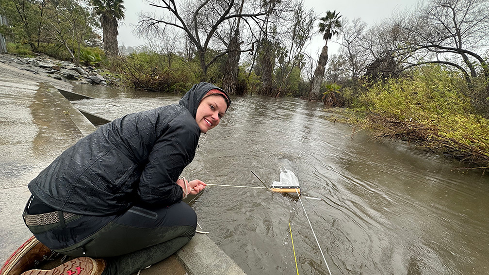 A young woman wearing raingear is kneeling on the cincrete bank of a river, holding a line connected to something submerged in the water. Palms, trees barren of leaves and other vegetation line the river in the distance.